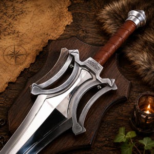 May include: A silver-toned metal sword featuring a wide, pointed blade and a complex, curved crossguard with engraved markings. The handle is wrapped in brown leather and finished with a rounded silver pommel, mounted on a dark wooden plaque.