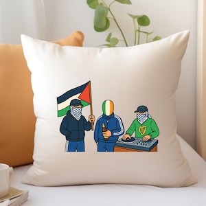 May include: Cream-coloured cushion featuring a cartoon illustration of three figures. One holds a Palestinian flag, another a beer, and the third is at a DJ setup. The figures wear tracksuits and face coverings.