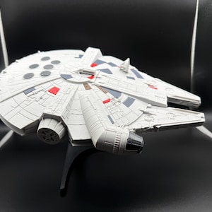 May include: A detailed model of the Millennium Falcon starship, predominantly white with gray and red accents. The model features intricate paneling and engine details, mounted on a black stand. A collectible item.