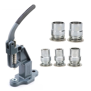 Op de afbeelding: Een grijze knopenmaker met een zwarte handgreep en verschillende zilveren knopenmaakcomponenten. De machine heeft een hendel en een veermechanisme. De componenten zijn cilindrisch met een cirkelvormige basis.