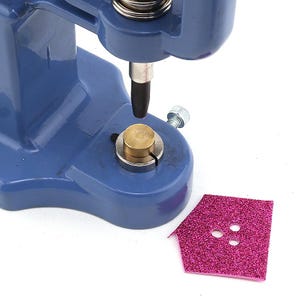 May include: A blue metal press with a brass base and a black tip, used for setting rivets or snaps. A pink glittery house-shaped piece of material with three holes is next to the press.