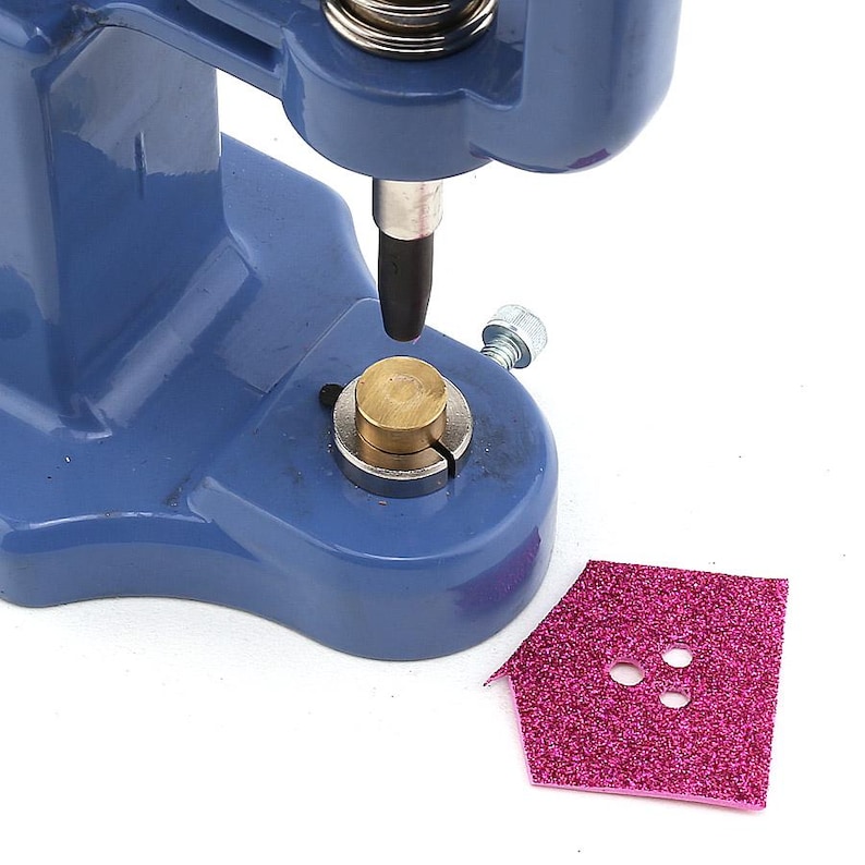 May include: A blue metal press with a brass base and a black tip, used for setting rivets or snaps. A pink glittery house-shaped piece of material with three holes is next to the press.