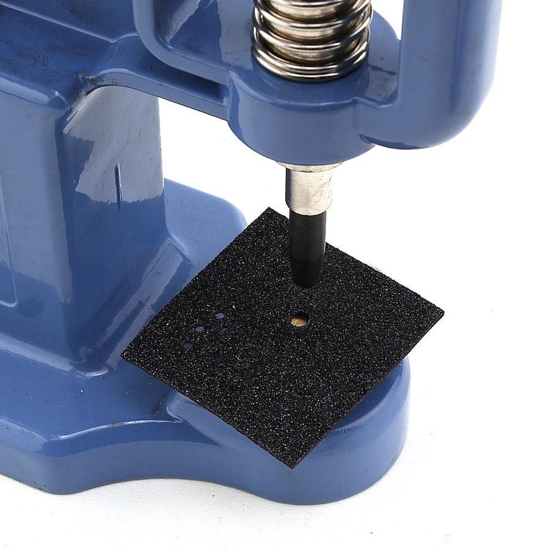 May include: A blue manual press machine with a silver spring and a black square pad. The machine has a metal rod extending downward, positioned over the pad. The pad has a hole in the center and several smaller holes.