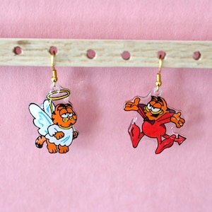 May include: A pair of dangle earrings featuring cartoon Garfield characters. One earring depicts Garfield as an angel with a halo and wings, the other as a devil in a red outfit. The earrings hang from gold-colored hooks.