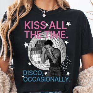 Op de afbeelding: Een zwart t-shirt met een grafische print van een grote zilveren discobal en een zwart-witfoto van een persoon die een bril rechtzet. De tekst luidt KISS ALL THE TIME in het roze en DISCO, OCCASIONALLY in het blauw, met witte sterren.
