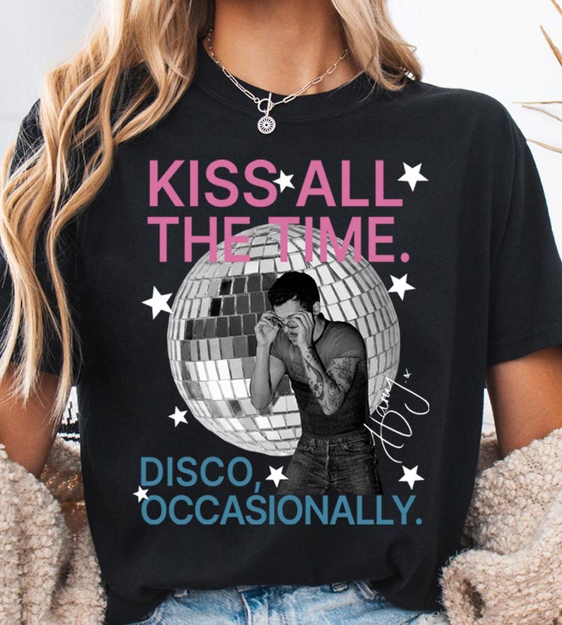 Op de afbeelding: Een zwart t-shirt met een grafische print van een grote zilveren discobal en een zwart-witfoto van een persoon die een bril rechtzet. De tekst luidt KISS ALL THE TIME in het roze en DISCO, OCCASIONALLY in het blauw, met witte sterren.