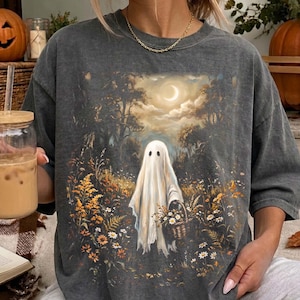 May include: A charcoal gray short-sleeve t-shirt featuring a graphic print of a white ghost standing in a field of wildflowers and tall grass. The ghost holds a woven basket filled with flowers under a night sky with a crescent moon and trees.