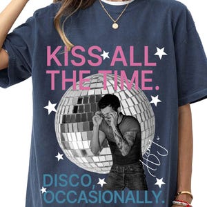 Op de afbeelding: Een marineblauw t-shirt met een grafische print van een discobal en een persoon die een bril rechtzet. De tekst luidt KISS ALL THE TIME in roze en DISCO, OCCASIONALLY in blauw, met witte sterren en een handtekening aan de rechterkant.