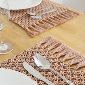 May include: A table setting with a woven, light brown placemat. A white plate, a spoon, and a knife rest on the mat. A wine glass is in the background. The placemat has a fringe. The setting is on a wooden table.
