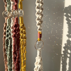 May include: Macrame plant hangers in various colors, including cream, yellow, and rust. Each hanger features a clear glass orb and gold-colored metal rings. The hangers are suspended, showcasing their intricate knotted designs.