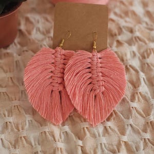 May include: A pair of handmade, coral-colored macrame earrings in a leaf design. The earrings are displayed on a brown card with gold-colored ear wires. The background includes a spool of coral-colored yarn and a textured, off-white fabric.