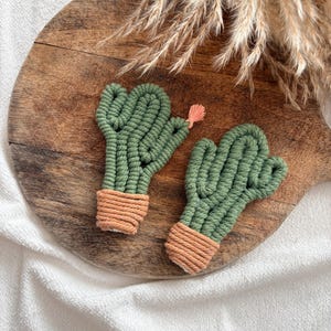 Pode incluir: Duas decorações de cactos de macramé feitas à mão. Os cactos são verdes com uma base castanha e uma pequena flor rosa. São exibidos numa tábua de cortar de madeira com erva seca no fundo. Um item de decoração para casa único.