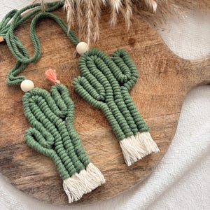 Peut inclure: Deux ornements cactus en macramé vert avec des franges blanches et des perles en bois, suspendus à une corde verte. La décoration cactus est présentée sur une planche à découper en bois, avec des plantes séchées en arrière-plan.