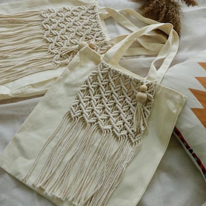 Peut inclure: Deux sacs fourre-tout en toile écrue avec des détails en macramé. Le macramé présente un motif géométrique et des franges. Des perles en bois ornent le haut du macramé. Les sacs ont de longues anses écrue.