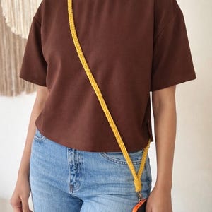 May include: A brown t-shirt and blue jeans are worn with a yellow braided phone strap. The phone case is orange. The strap is worn across the body. The outfit is casual and simple.