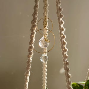 May include: A cream macrame plant hanger with gold metal detailing. The design includes two gold rings, a leaf and butterfly charm, and two clear crystal prisms. The hanger is designed to hold a potted plant, adding a decorative touch.