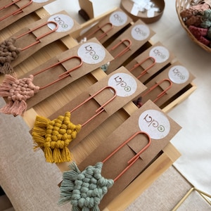 May include: A collection of large copper-colored metal paper clips, each featuring a small macrame knot design in various colors including mustard, sage green, pink, and brown. Each clip is attached to a rectangular card with a circular 'eda' logo.