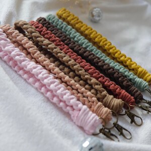 May include: A collection of macrame wrist lanyards in various colors, including pink, tan, brown, rust, green, and yellow. Each lanyard features a woven design and a bronze-colored clasp for attaching keys or other small items.