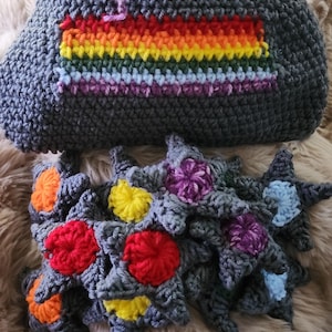 May include: A crocheted gray bag with a rainbow stripe design and a set of star-shaped memory game pieces. The stars are gray with colorful flower centers in red, orange, yellow, purple, and blue.