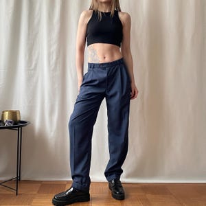May include: Navy blue pleated trousers with a high waist. The pants have a relaxed fit and are paired with a black crop top and black platform loafers. The trousers are made of a smooth fabric.