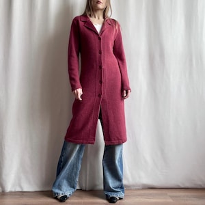 Può includere: Un cardigan lungo fino al ginocchio, a maniche lunghe e con chiusura a bottoni, di colore bordeaux intenso, caratterizzato da un colletto e una trama in maglia. Il capo è abbinato a jeans larghi in denim blu, dal taglio ampio e lavaggio chiaro.