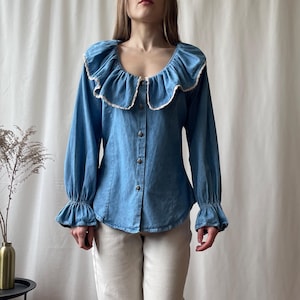 Könnte beinhalten: Eine hellblaue Jeansbluse mit Rüschenkragen und Manschetten. Das langärmlige Hemd hat eine Knopfleiste vorne und eine weiße Spitzenborte am Kragen. Die Bluse ist im Vintage-Stil gehalten.