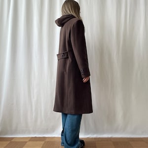 Peut inclure: Un long manteau marron foncé avec une capuche et des boutons. Le manteau est porté sur un jean bleu. Le manteau a une coupe ajustée et semble être fait d'un tissu en laine ou en mélange de laine. Le manteau est de style classique.
