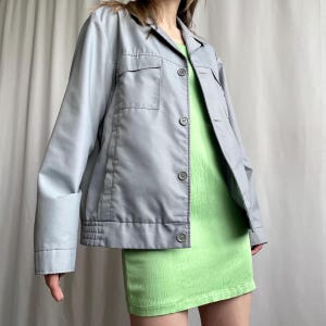 May include: A pale blue jacket with two front pockets and button fastenings. The jacket is worn over a short, lime green dress. The sleeves are cuffed at the wrists. The outfit is displayed against a neutral background.