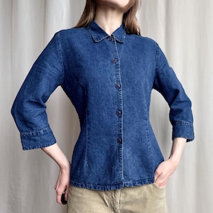 Pode incluir: Uma camisa de ganga azul escura com gola clássica e fecho de botões. A camisa tem mangas três quartos e uma silhueta ajustada. O tecido parece ser ganga de peso médio, adequada para várias ocasiões.