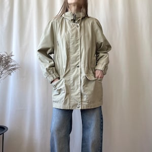 May include: A beige jacket with a high collar, zip, and popper fastenings. The jacket features two front pockets and is worn with blue jeans. The sleeves have elasticated cuffs. The model is standing in front of a white curtain.