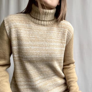 May include: A beige and white striped turtleneck jumper. The jumper has long sleeves and a ribbed turtleneck collar. The knitwear is a blend of beige and white, creating a textured, horizontal striped pattern. The sleeves are a solid beige colour.