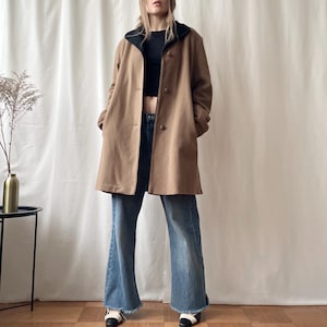 Puede incluir: Un abrigo color canela de largo medio con cuello negro y tres botones frontales, usado sobre un top corto negro y jeans azules con dobladillos deshilachados. El abrigo presenta mangas largas y una silueta recta, ofreciendo una capa exterior neutra.