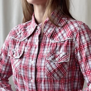 May include: A long-sleeve button-down shirt featuring a red, white, and blue plaid pattern with small floral accents. The shirt includes a pointed collar, two chest pockets with buttoned flaps, and white buttons running down the front placket.