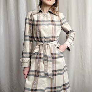 May include: A long-sleeved trench coat featuring a beige, brown, and dark grey plaid pattern. The coat includes a pointed collar, a front button closure, two side pockets, and a matching fabric tie belt at the waist with buttoned sleeve cuffs.