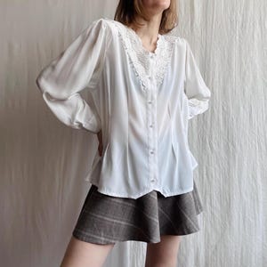 May include: A white blouse featuring a lace collar and pearl buttons. The long-sleeved top has a slightly puffed shoulder design. The blouse is paired with a brown plaid skirt. The outfit is set against a white backdrop.