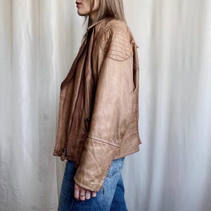 May include: A tan leather jacket with a classic design. The jacket features a notched collar, a zip closure, and quilted shoulder detailing. The sleeves have zips at the cuffs. The jacket is worn with blue jeans.