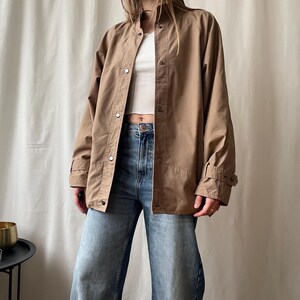 Op de afbeelding: Een bruine jas met een opstaande kraag en drukknoopsluitingen. De jas heeft lange mouwen met verstelbare manchetten en wordt gecombineerd met een blauwe jeans en een witte top. De jas heeft een neutrale kleur en lijkt van een lichte stof te zijn.