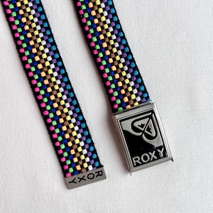 Puede incluir: Un cinturón Roxy colorido con un patrón tejido de pequeños cuadrados en varios colores, incluyendo rosa, amarillo, azul y verde. El cinturón tiene una hebilla de metal plateado con el logo de Roxy.
