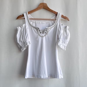 Puede incluir: Top blanco con hombros descubiertos y mangas cortas con volantes. El escote presenta un ribete plateado y un detalle de cordones. La parte superior está colgada de una percha de madera sobre un fondo blanco liso.