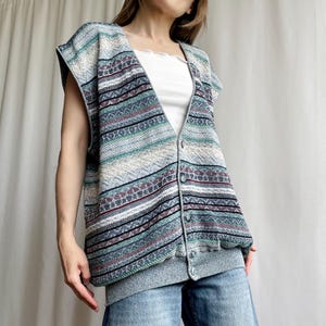 May include: A sleeveless, button-down vest with a geometric pattern in shades of blue, green, and burgundy. The vest is worn over a white top and paired with denim shorts. The knitted fabric has a textured appearance.