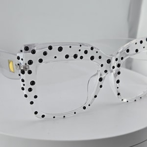 Pode incluir: Óculos transparentes com armação quadrada, adornados com bolinhas pretas nas bordas. Os óculos têm armação e lentes transparentes, oferecendo um visual moderno e elegante. O design é simples, mas atraente.
