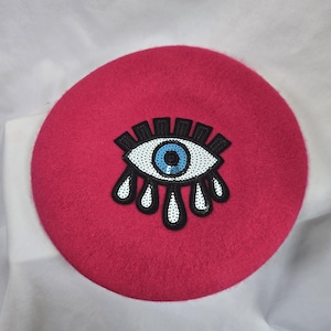 May include: A vibrant pink beret featuring an embroidered eye motif. The eye design includes a blue iris, white detailing, and black outlines. The beret is circular and made from a soft fabric.