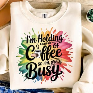 May include: Cream-colored sweatshirt with a colorful paint splatter design. The graphic reads "I'm Holding a cup of coffee so I'm pretty busy" in black script and block letters. A hand holds a coffee cup.