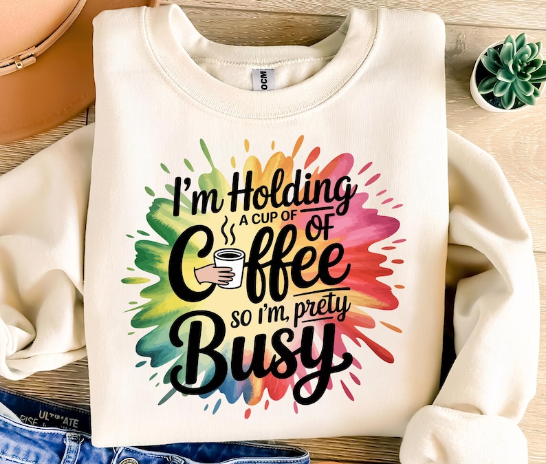 May include: Cream-colored sweatshirt with a colorful paint splatter design. The graphic reads "I'm Holding a cup of coffee so I'm pretty busy" in black script and block letters. A hand holds a coffee cup.