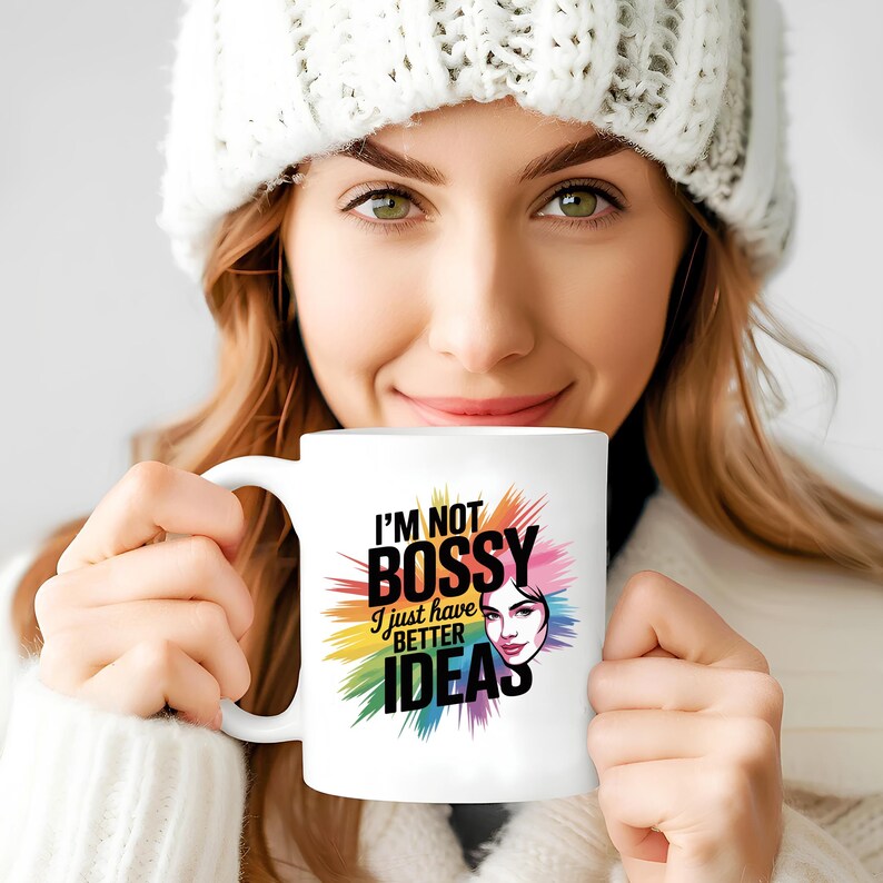 May include: White ceramic mug with the text "I'M NOT BOSSY I just have BETTER IDEAS" in black, rainbow, and pink colors. A woman's face is also featured on the mug. The mug is held by a person wearing a white knit hat.