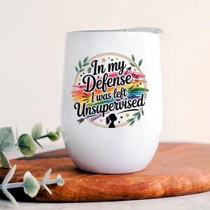 May include: White insulated wine tumbler with a lid. The tumbler features the text "In my Defense I was left Unsupervised" in a colorful design with a rainbow and floral accents. The tumbler is on a wooden surface.