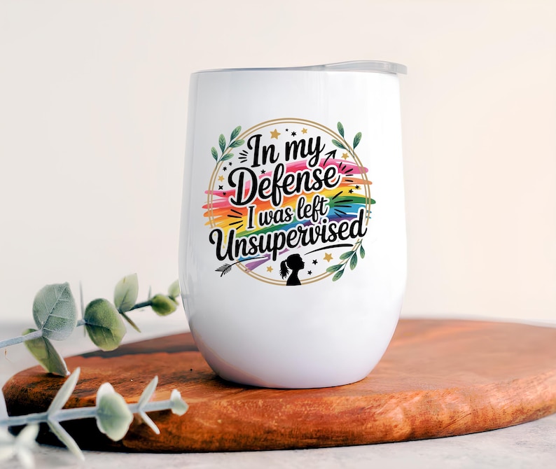 May include: White insulated wine tumbler with a lid. The tumbler features the text "In my Defense I was left Unsupervised" in a colorful design with a rainbow and floral accents. The tumbler is on a wooden surface.