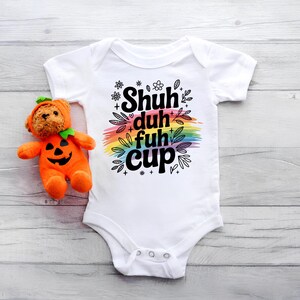 May include: White baby onesie with the text "Shuh duh fuh cup" in black, over a rainbow-colored brushstroke. A plush orange pumpkin toy with a bear face is next to the onesie.