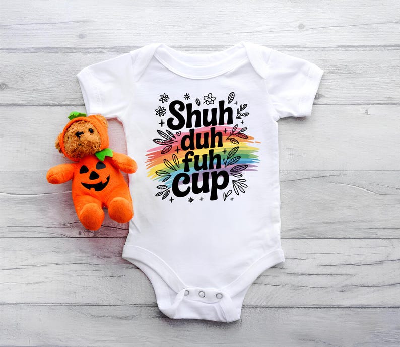 May include: White baby onesie with the text "Shuh duh fuh cup" in black, over a rainbow-colored brushstroke. A plush orange pumpkin toy with a bear face is next to the onesie.