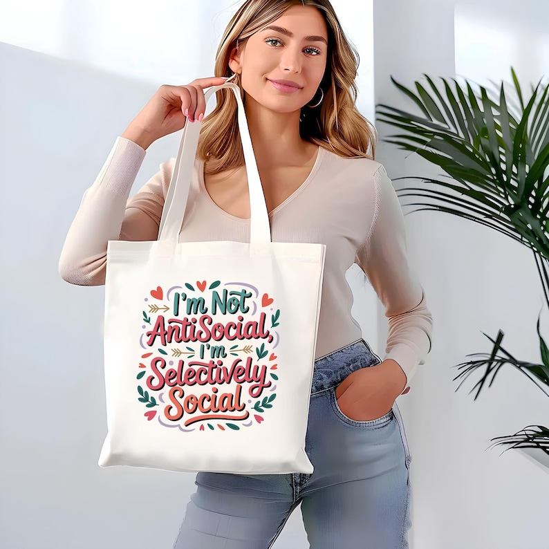 May include: A white tote bag with the text "I'm Not AntiSocial, I'm Selectively Social" in a colorful, decorative font. The bag has long handles and is held up by a person. The design includes hearts and leaves.
